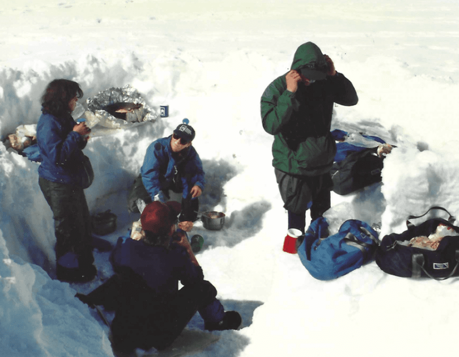 People gathered on snowy ground, some sitting and some standing with gear.
