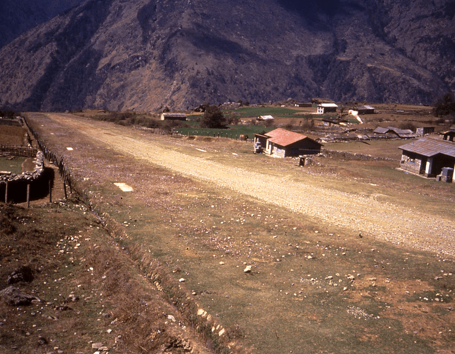 A rural dirt airstrip with a small village and mountains in the background.