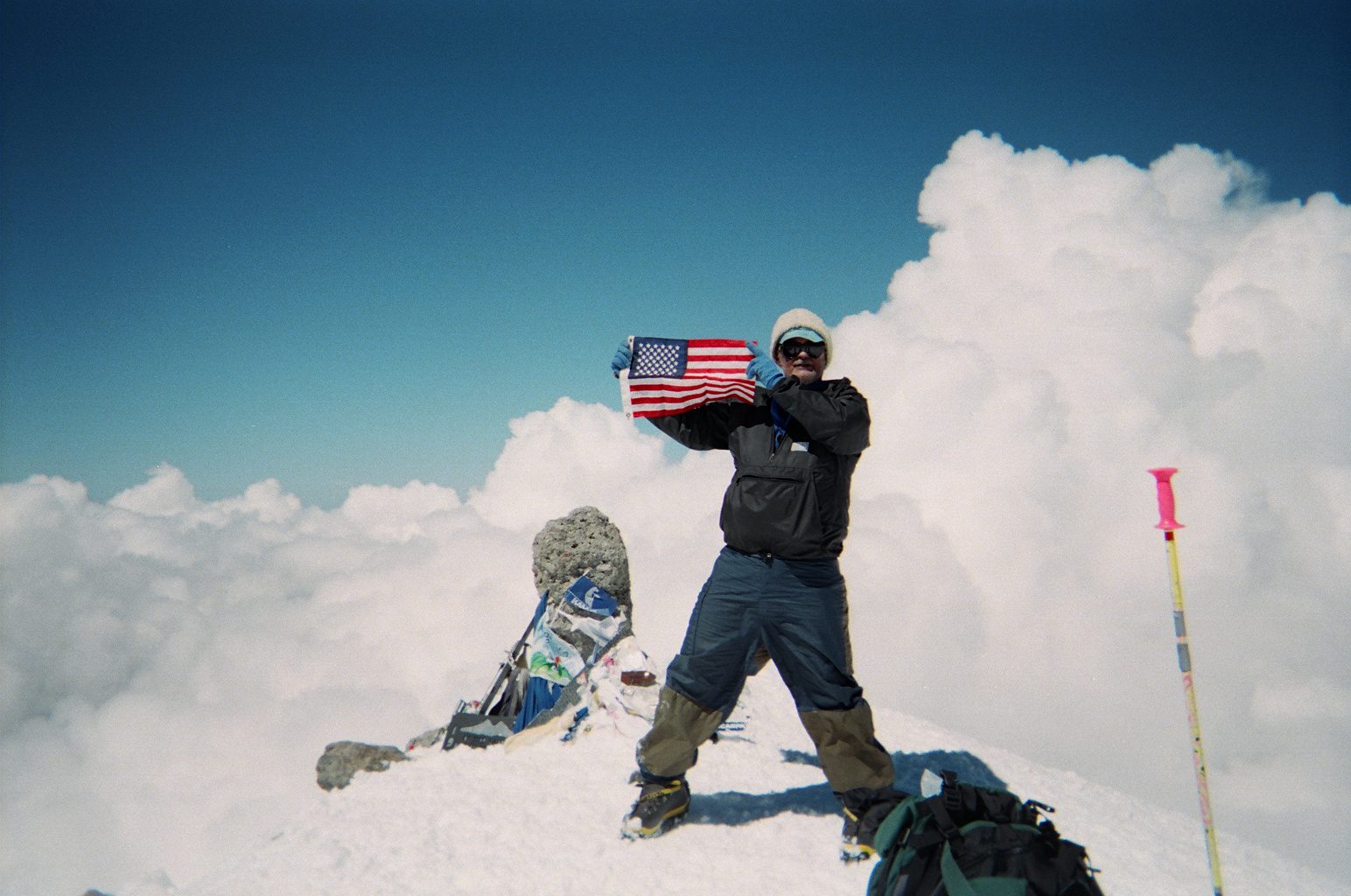 Climber on snowy peak holding an American flag with gear and rope nearby.