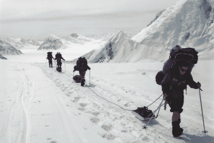 Group trekking through snowy mountainous terrain in harsh winter conditions.
