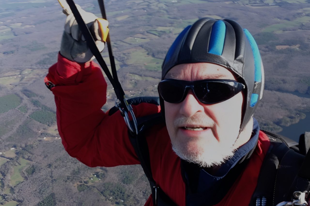 Man in helmet and sunglasses paragliding over countryside.