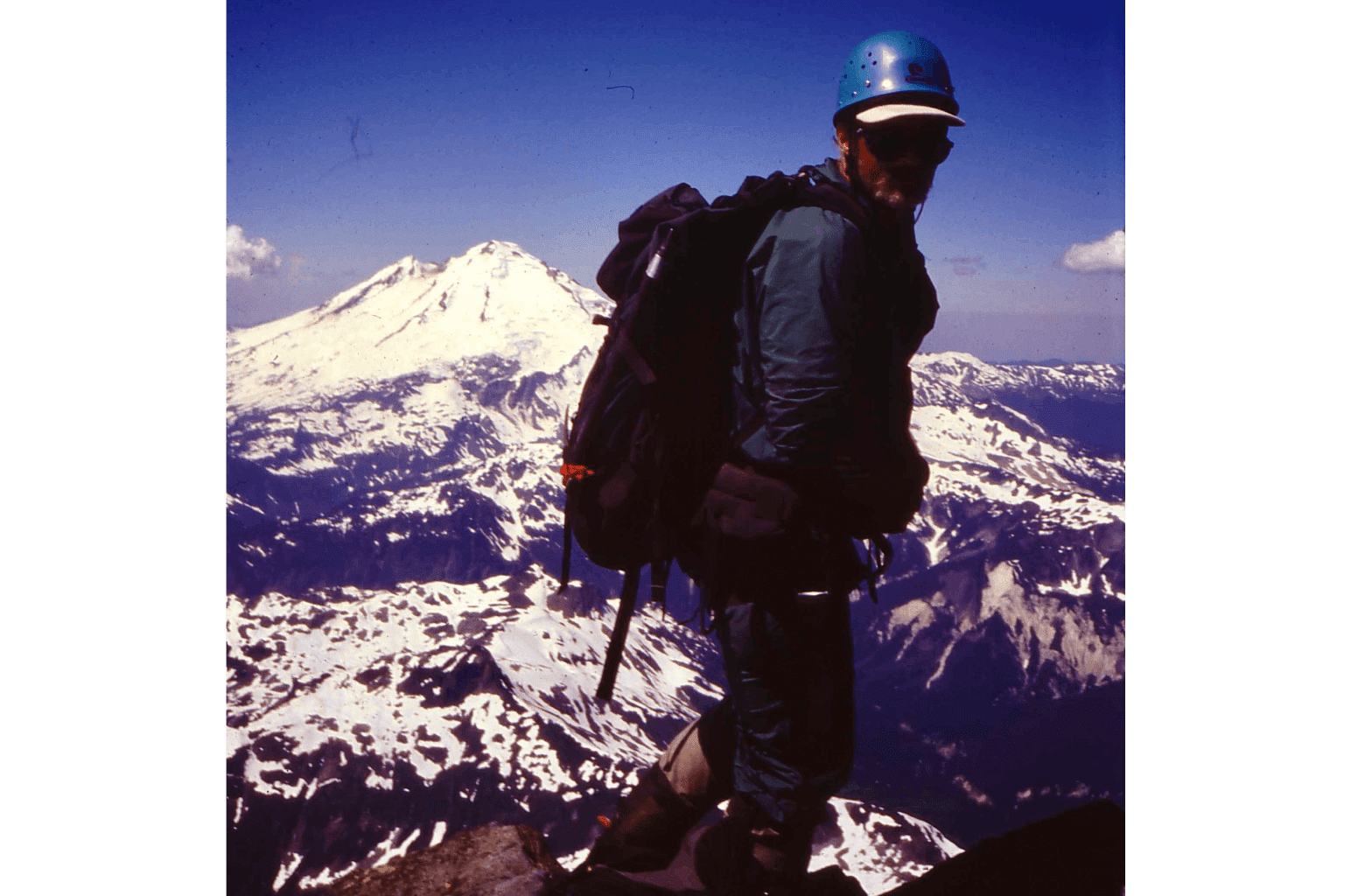 Hiker with backpack overlooking snow-capped mountains under clear blue sky.
